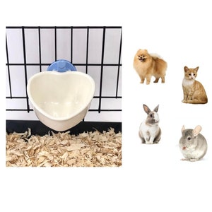 May include: A white ceramic heart-shaped pet bowl with a blue suction cup attached to the side. The bowl is mounted on a black wire cage with wood shavings on the bottom.