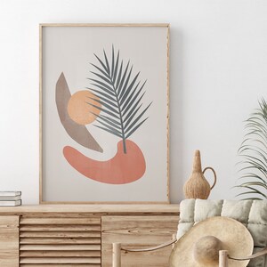 K&ouml;nnte beinhalten: Ein gerahmter Druck mit einem abstrakten Design, das ein stilisiertes Palmblatt, einen pfirsichfarbenen Kreis und eine braune Sichelform auf einem beigefarbenen Hintergrund zeigt.