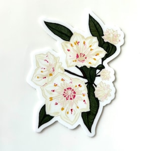 May include: A white flower sticker with pink and yellow accents. The flower has five petals and is surrounded by green leaves.