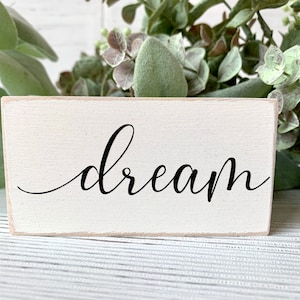 May include: White wooden sign with the word "dream" written in black cursive script.