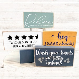 May include: Five wooden signs with various sayings. The top sign is light blue with white text that reads "Relax Let Your Troubles Melt Away". The second sign is white with black text that reads "Would Poop Here Again!" and has five black stars. The third sign is yellow with black text that reads "hey sweet cheeks". The fourth sign is dark gray with white text that reads "Wash your hands ya filthy animal". The fifth sign is dark gray and is blank.