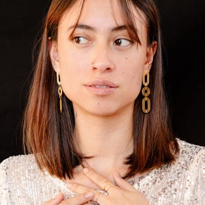 Peut inclure: Une femme portant des boucles d'oreilles en or avec un motif géométrique. Les boucles d'oreilles sont composées de plusieurs formes ovales empilées les unes sur les autres.