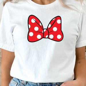 May include: White t-shirt featuring a red bow with white polka dots. The bow is outlined in black. The shirt is a crew neck and is made of a soft material. The shirt is paired with blue denim jeans.