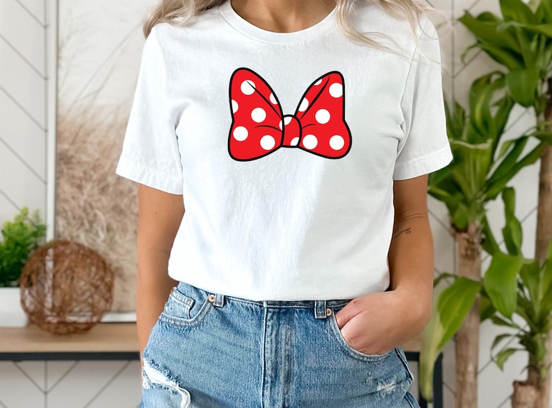 May include: White t-shirt featuring a red bow with white polka dots. The bow is outlined in black. The shirt is a crew neck and is made of a soft material. The shirt is paired with blue denim jeans.