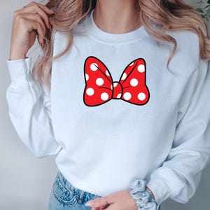 May include: A white sweatshirt featuring a large red bow with white polka dots. The bow has a black outline. The sweatshirt has long sleeves and a crew neck. The design is centered on the chest.