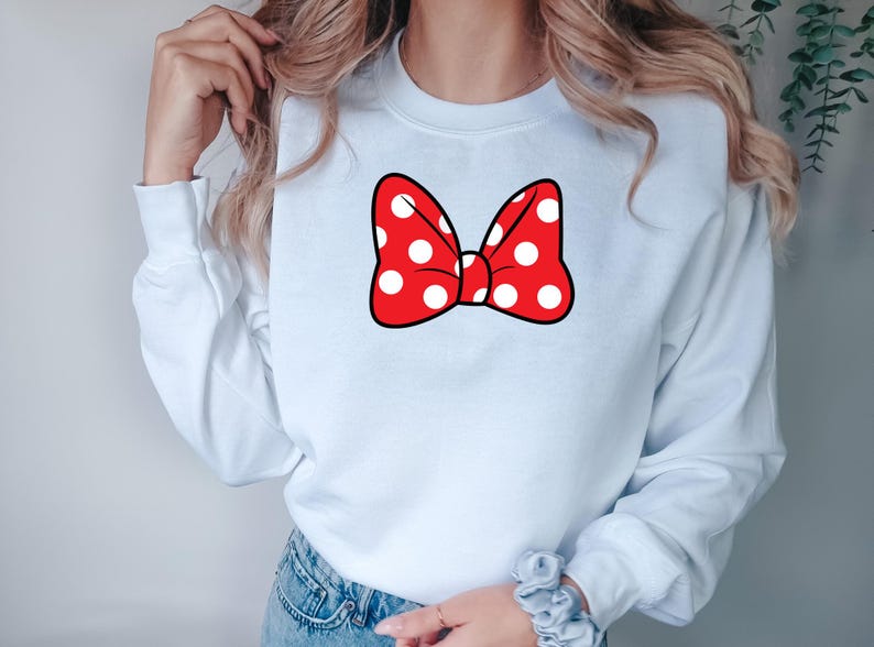 May include: A white sweatshirt featuring a large red bow with white polka dots. The bow has a black outline. The sweatshirt has long sleeves and a crew neck. The design is centered on the chest.