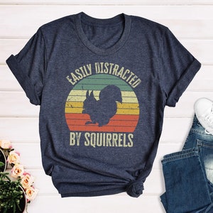 May include: A navy blue t-shirt with a vintage-style graphic of a squirrel in front of a sunset. The text "Easily Distracted By Squirrels" is printed on the shirt.