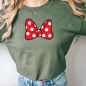 May include: An olive green sweatshirt featuring a large red bow with white polka dots. The bow is centered on the chest. The sweatshirt is made of a soft material and has long sleeves.