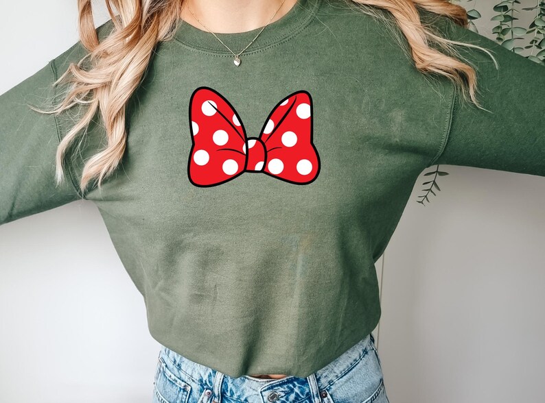 May include: An olive green sweatshirt featuring a large red bow with white polka dots. The bow is centered on the chest. The sweatshirt is made of a soft material and has long sleeves.
