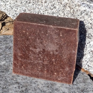 May include: A rectangular bar of brown soap with a speckled appearance. The soap is sitting on a gray, textured surface.