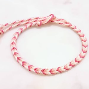 May include: A pink and white braided friendship bracelet. The bracelet is made of thin string and has a chevron pattern.