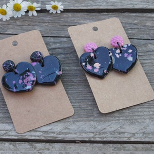 May include: Two pairs of black heart-shaped earrings with pink and white speckles. The earrings are on brown card stock.