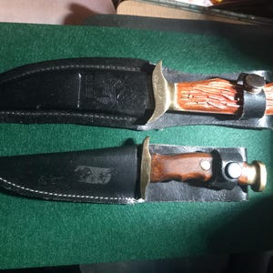 May include: Two hunting knives with black leather sheaths. The knives have wooden handles and brass bolsters. One handle is a reddish-brown color, and the other is a darker brown. The sheaths are embossed with designs and have white stitching.