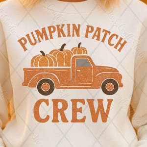 May include: A cream-coloured sweatshirt with the words "PUMPKIN PATCH CREW" in orange. The design shows an orange truck loaded with pumpkins. The sweatshirt is a crew neck style with long sleeves. The model is wearing blue jeans.