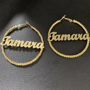 Puede incluir: Un par de pendientes de aro dorados con el nombre "Tamara" escrito en ellos.