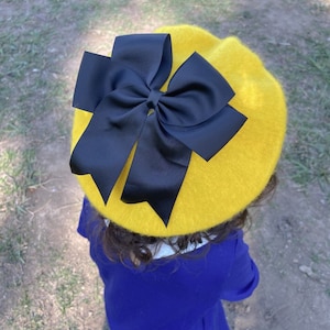 May include: A yellow beret with a large black bow on top. The beret is made of a soft, fuzzy material.