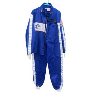 May include: A blue racing suit with white stripes down the sides. The jacket has a zipper closure, a small American flag patch, and a logo on the chest. The pants have elastic cuffs at the ankles.
