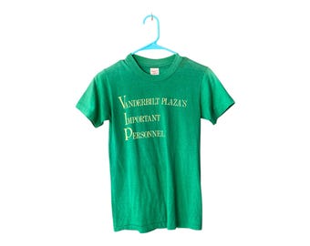 Könnte beinhalten: Grünes T-Shirt mit kurzen Ärmeln und dem Aufdruck "Vanderbilt Plaza's Important Personnel" in Gelb.