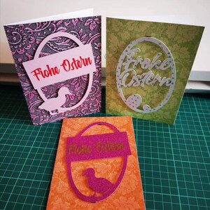 Pu&ograve; includere: Tre biglietti di Pasqua fatti a mano con diversi design. Il primo biglietto ha uno sfondo rosa con un ritaglio ovale bianco con il testo "Frohe Ostern" in rosso. Il secondo biglietto ha uno sfondo verde con un ritaglio ovale bianco con il testo "Frohe Ostern" in bianco. Il terzo biglietto ha uno sfondo arancione con un ritaglio ovale rosa con il testo "Frohe Ostern" in bianco.