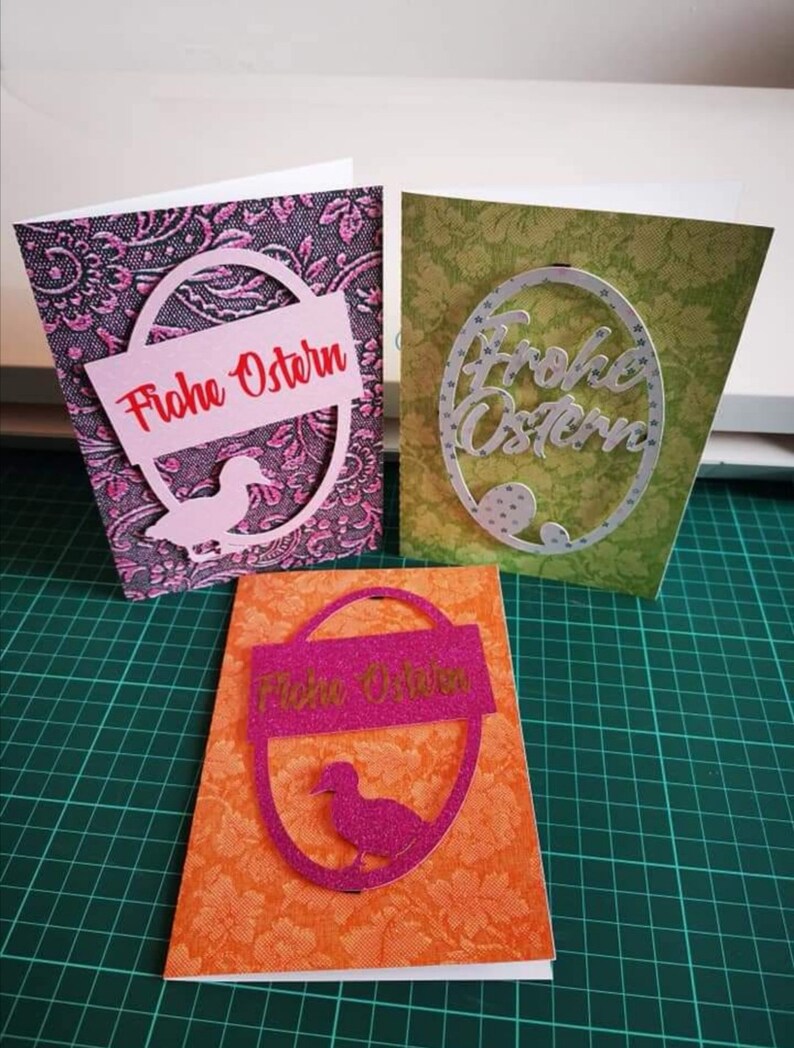 Pu&ograve; includere: Tre biglietti di Pasqua fatti a mano con diversi design. Il primo biglietto ha uno sfondo rosa con un ritaglio ovale bianco con il testo "Frohe Ostern" in rosso. Il secondo biglietto ha uno sfondo verde con un ritaglio ovale bianco con il testo "Frohe Ostern" in bianco. Il terzo biglietto ha uno sfondo arancione con un ritaglio ovale rosa con il testo "Frohe Ostern" in bianco.