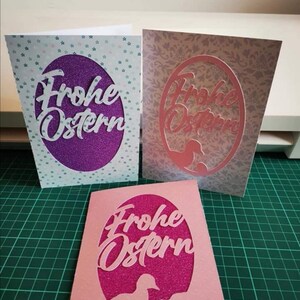 Pu&ograve; includere: Tre biglietti di Pasqua fatti a mano con sfondo viola, rosa e bianco. Ogni biglietto presenta un ovale ritagliato con il testo "Frohe Ostern" in bianco, rosa e bianco. Gli ovali sono decorati con glitter.