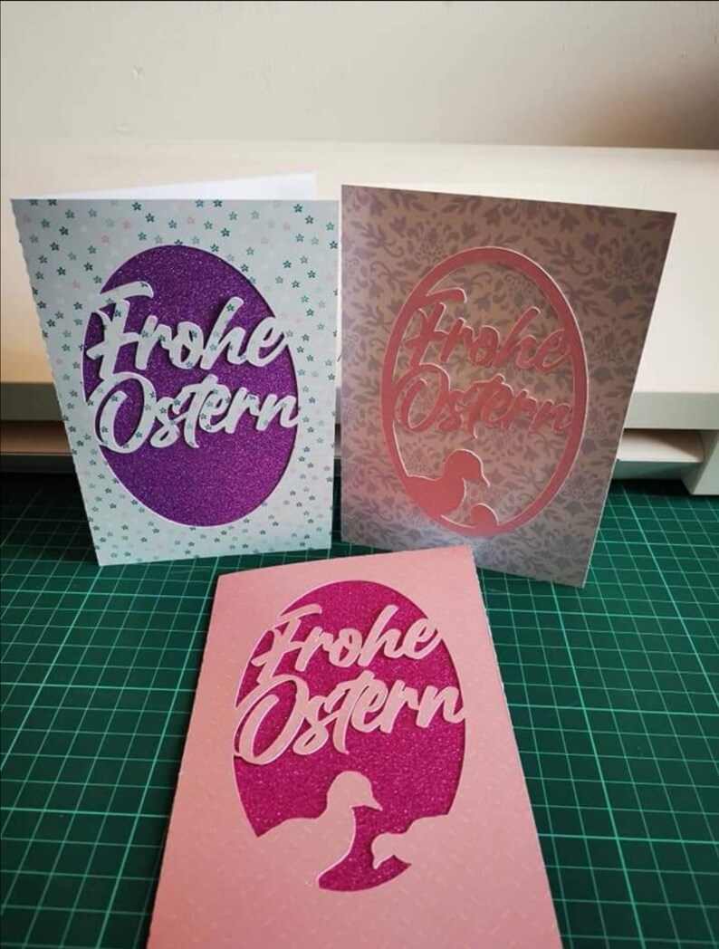 Pu&ograve; includere: Tre biglietti di Pasqua fatti a mano con sfondo viola, rosa e bianco. Ogni biglietto presenta un ovale ritagliato con il testo "Frohe Ostern" in bianco, rosa e bianco. Gli ovali sono decorati con glitter.