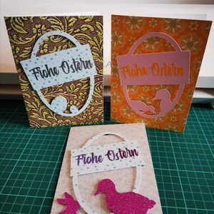 Pu&ograve; includere: Tre biglietti di Pasqua fatti a mano con diversi design. Ogni biglietto presenta un ovale bianco con il testo "Frohe Ostern" in lettere viola. Il primo biglietto ha uno sfondo marrone con un motivo floreale dorato e verde. Il secondo biglietto ha uno sfondo arancione con un motivo floreale giallo. Il terzo biglietto ha uno sfondo marrone chiaro con un ovale bianco, un'anatra glitterata rosa e un coniglietto glitterato rosa.