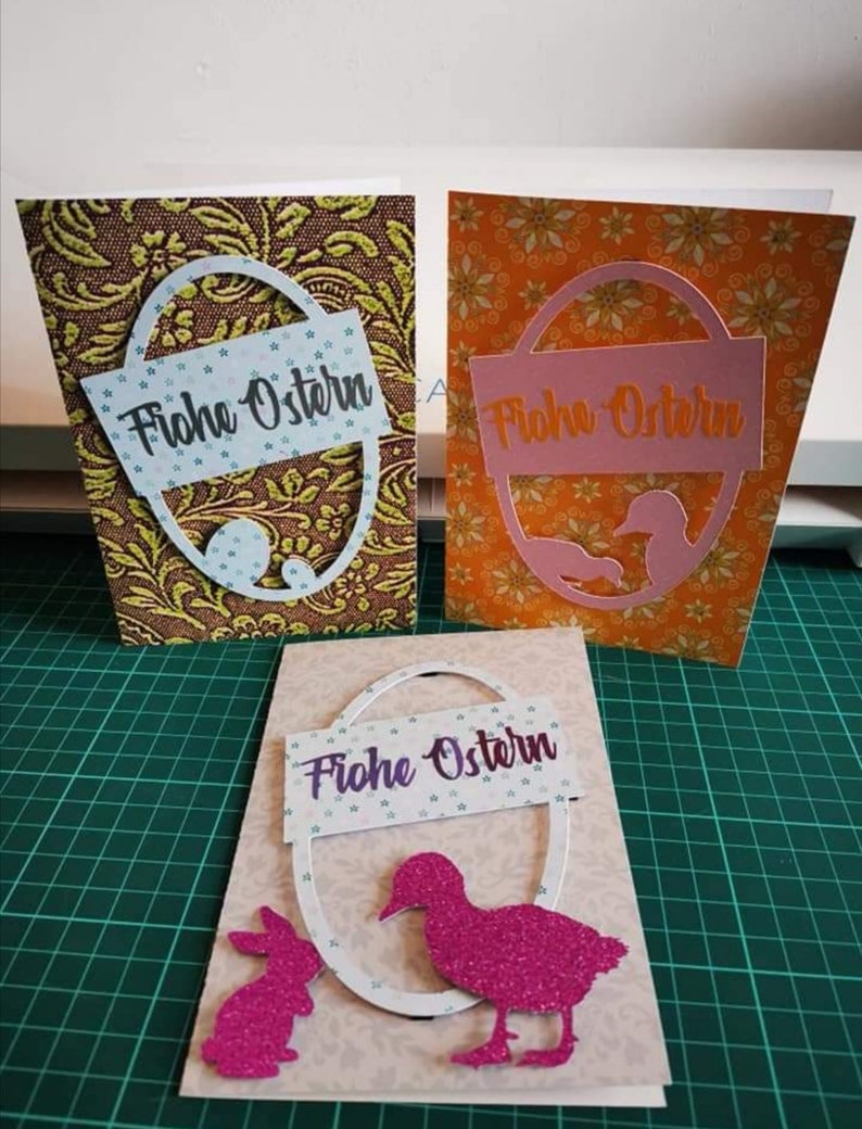 Pu&ograve; includere: Tre biglietti di Pasqua fatti a mano con diversi design. Ogni biglietto presenta un ovale bianco con il testo "Frohe Ostern" in lettere viola. Il primo biglietto ha uno sfondo marrone con un motivo floreale dorato e verde. Il secondo biglietto ha uno sfondo arancione con un motivo floreale giallo. Il terzo biglietto ha uno sfondo marrone chiaro con un ovale bianco, un'anatra glitterata rosa e un coniglietto glitterato rosa.