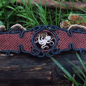 May include: A macrame bracelet with an octopus design. The bracelet has a reddish-brown centre with dark blue trim and a matching cord. The octopus is a light silver colour. The bracelet is displayed on a weathered log.