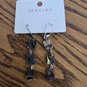 May include: A pair of drop earrings featuring three rectangular abalone shell beads each, suspended from silver-toned metal lever-back hooks. The earrings are attached to a white display card with the word JEWELRY printed in black at the top.