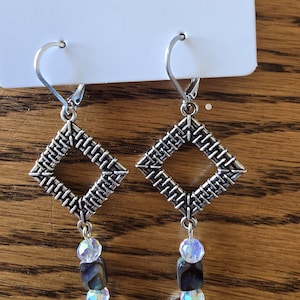 May include: Silver-toned earrings with a woven diamond-shaped top, dangling beads, and a peace sign charm. The earrings have a lever-back closure. The word "JEWELRY" is printed at the top.