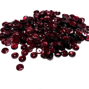 May include: A pile of dark red sequins, each with a flat, round shape and a small hole in the center. The sequins are scattered against a white background, creating a textured appearance. These shiny embellishments are ideal for crafts and sewing projects.