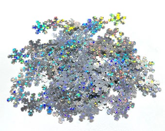 May include: A pile of silver snowflake-shaped confetti with a holographic, iridescent finish. The confetti reflects a spectrum of colors, including blue, green, and yellow, against a white background. Ideal for holiday crafts and decorations.