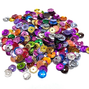 May include: A large pile of colorful iridescent sequins in various shades of purple, pink, blue, green, gold, silver, and red. The sequins are scattered on a white background.