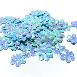 May include: A pile of iridescent blue flower-shaped sequins. The sequins have a light blue color with a subtle iridescent sheen.