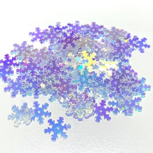 May include: A pile of iridescent blue snowflake-shaped sequins. The sequins are scattered on a white surface.