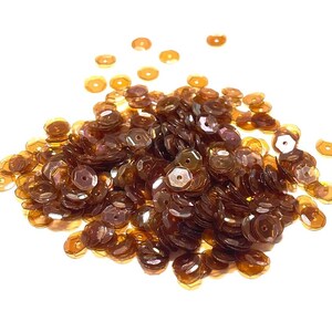 May include: A pile of brown sequins with a metallic sheen, scattered against a white background. The sequins are round with a central hole, reflecting light. Ideal for embellishments in crafts and sewing projects.
