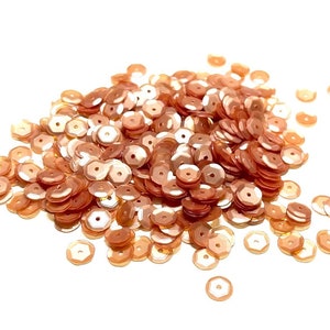 May include: A pile of peach-colored sequins, scattered on a white background. The sequins are small and round, with a slightly iridescent finish.