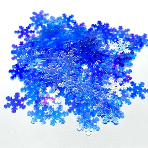 May include: A pile of iridescent blue snowflake-shaped glitter. The glitter is scattered on a white surface.