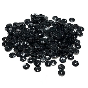 May include: A large pile of black sequins, scattered on a white background. The sequins are round and have a slightly raised center.