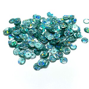 May include: A large pile of teal iridescent sequins. The sequins are small and round with a flat back.