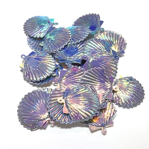 May include: A collection of iridescent seashell-shaped charms. The charms are a mix of blue, purple, and silver, with a shimmering, holographic effect. Each charm has a small loop for attaching to jewelry or other crafts. The charms are scattered on a white surface.