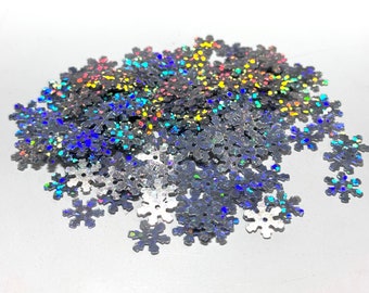 May include: A pile of silver snowflake-shaped confetti with a holographic, iridescent finish. The confetti is scattered, with varying sizes and shapes. The reflective surface catches and refracts light, creating a shimmering effect. Ideal for crafts and decorations.