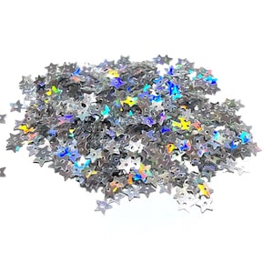 May include: A pile of iridescent silver star-shaped sequins. The sequins are scattered on a white background.