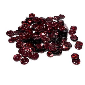 May include: A pile of maroon sequins, scattered on a white background. The sequins are round and have a slightly iridescent finish.