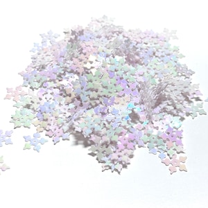 May include: A pile of iridescent snowflake-shaped sequins. The sequins are in shades of white, pink, green, and purple. The sequins are scattered on a white surface.