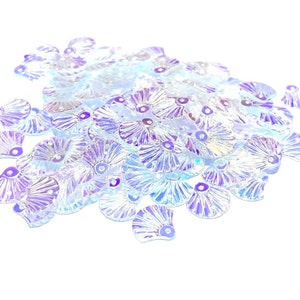 May include: A pile of iridescent, clear plastic seashell-shaped sequins. The sequins have a light purple hue and are scattered on a white background.