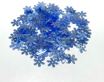 May include: A pile of blue iridescent snowflake-shaped sequins. The sequins are scattered on a white surface.