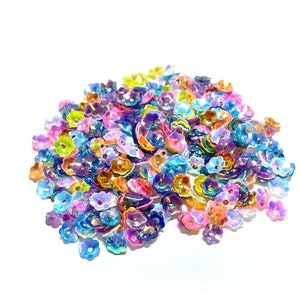 May include: A large pile of iridescent flower-shaped sequins in a variety of colors, including pink, blue, purple, and orange.