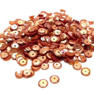 May include: A pile of copper-colored hexagonal sequins, scattered on a white background.